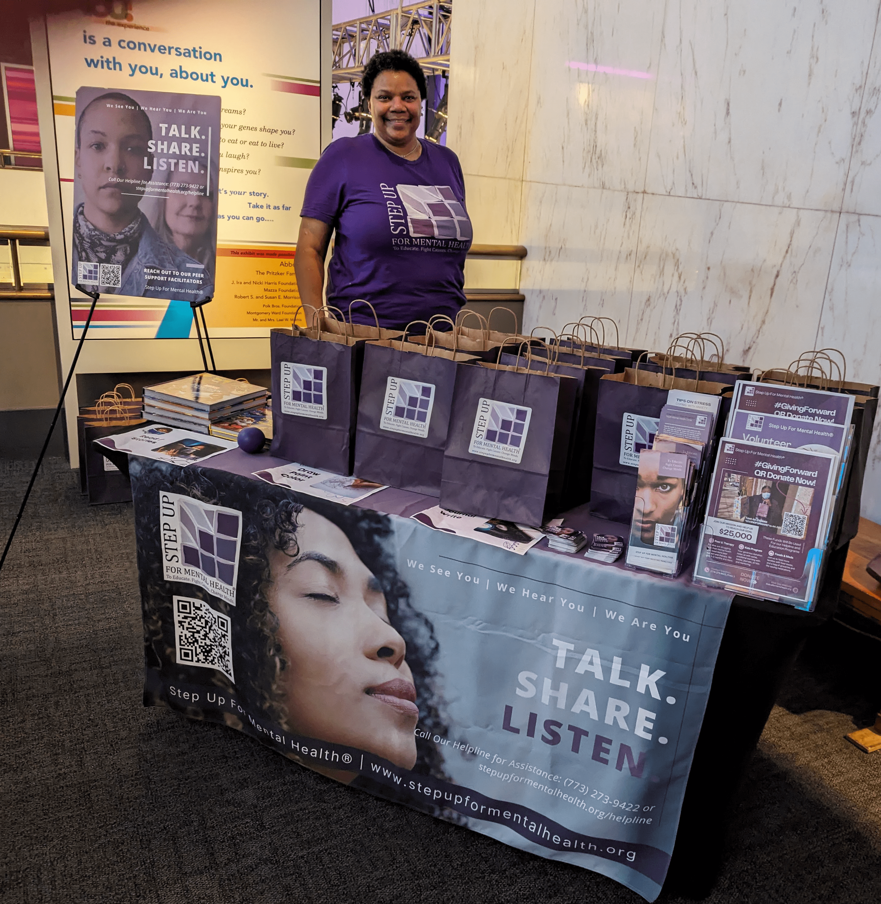 A person in a purple shirt standing at a conference booth with goodie bags that say "Step Up for Mental Health"